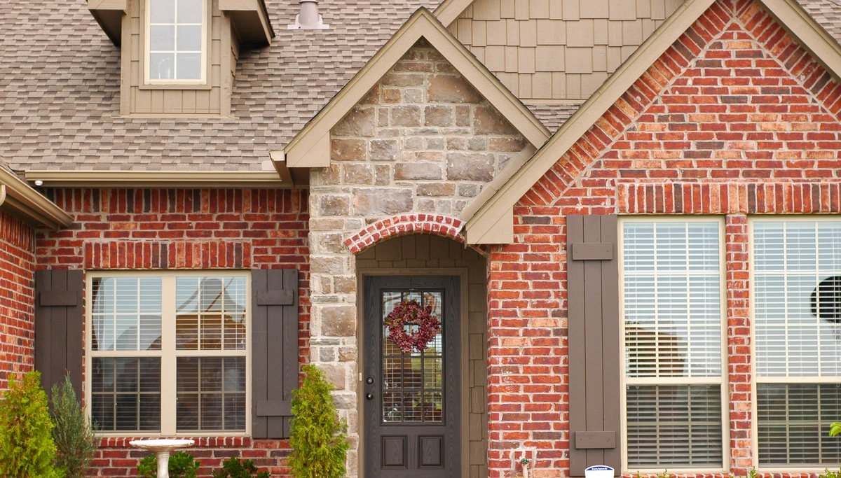 The suburban house boasts a red brick facade complemented by stone entryway and gray front door, showcasing harmonious house siding colors. Two large windows with dark shutters flank the entrance beneath a brown gabled roof. A wreath decorates the door, while small shrubs adorn the front.