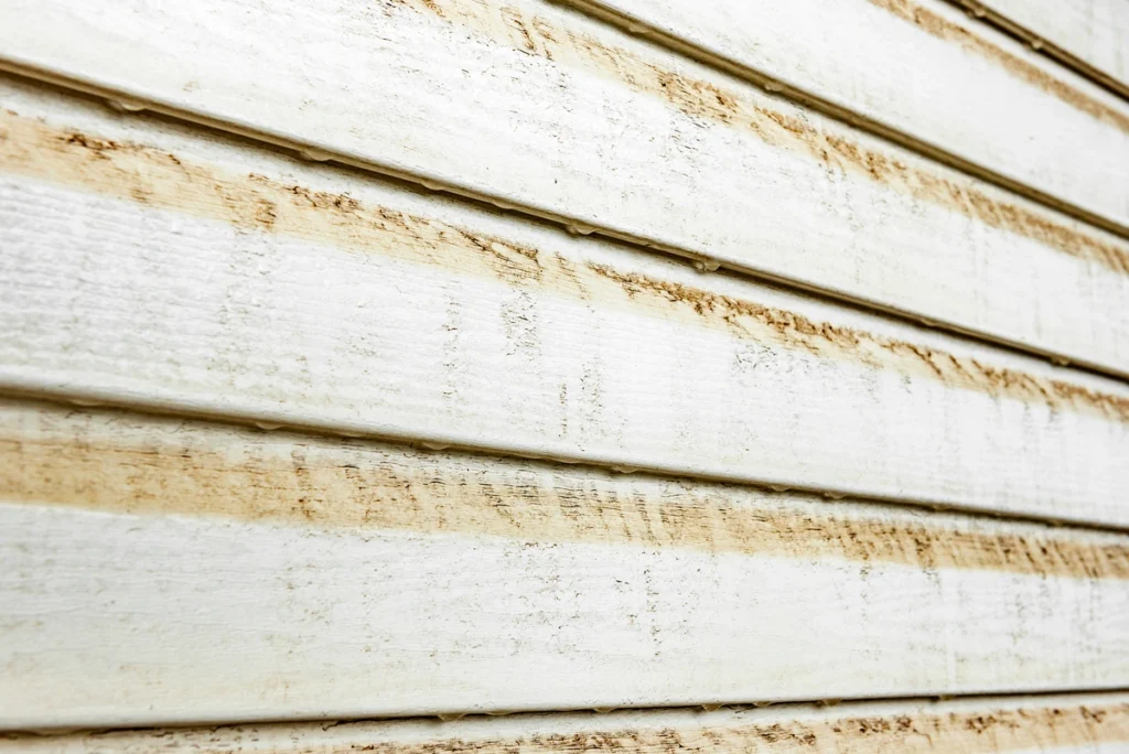 Close-up of weathered white siding on an exterior wall, showing horizontal panels with visible dirt and wear. The texture is varied, highlighting the aged and rustic appearance of the surface.