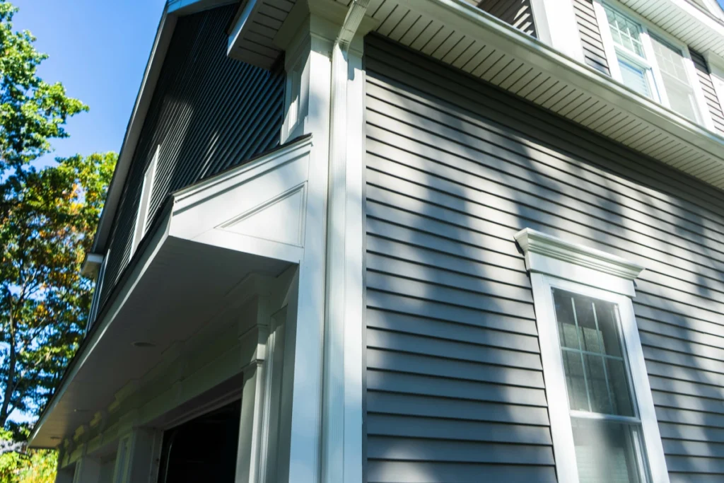 Close-up of a modern house's exterior highlighting tasteful house siding colors with grey horizontal panels and white trim. The structure features a window and an open garage, with sunlight casting shadows. A hint of blue sky and green foliage complements the design.