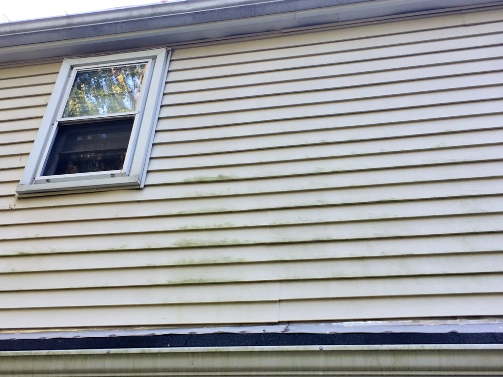 Exterior view of a house showing beige horizontal vinyl siding with visible discoloration and stains. A single window with a white frame is located in the upper left section of the wall.