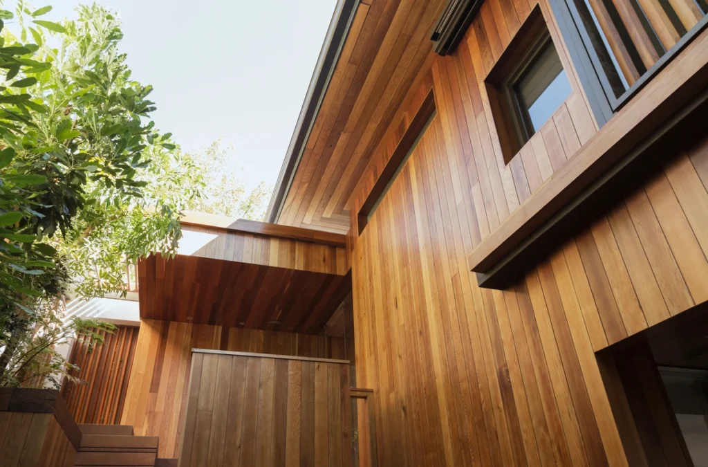 The modern wooden house facade features sleek panels and a sophisticated design, showcasing siding types that enhance its elegance. A small upper balcony and a set of stairs are highlighted, while lush green trees partially surround the building, complementing its natural wood texture.