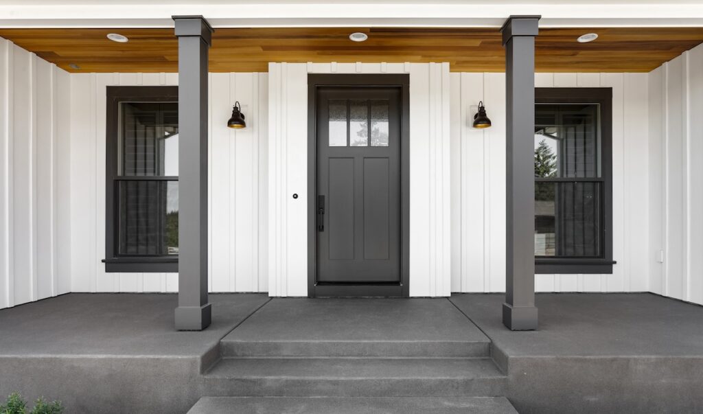 A modern house entrance with a gray door flanked by two large windows. It features white vertical siding, a wooden porch ceiling, and two black column supports. Steps lead up to the porch, and two wall-mounted lights are on either side of the door.