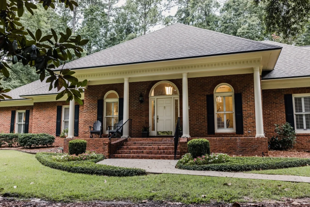A brick house with a large porch featuring white columns and a central white door. Arched windows with dark shutters flank the entrance, complementing the best siding for house elegance. The front yard showcases manicured shrubs and a curved walkway, surrounded by lush greenery.