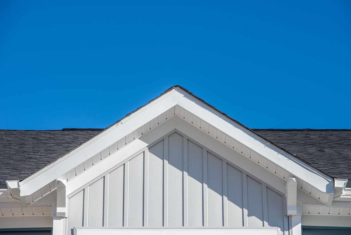 A close-up view of a house's gable roof with white horizontal siding and a dark shingled roof against a clear blue sky. The triangular shape of the roof is prominent, with clean architectural lines.