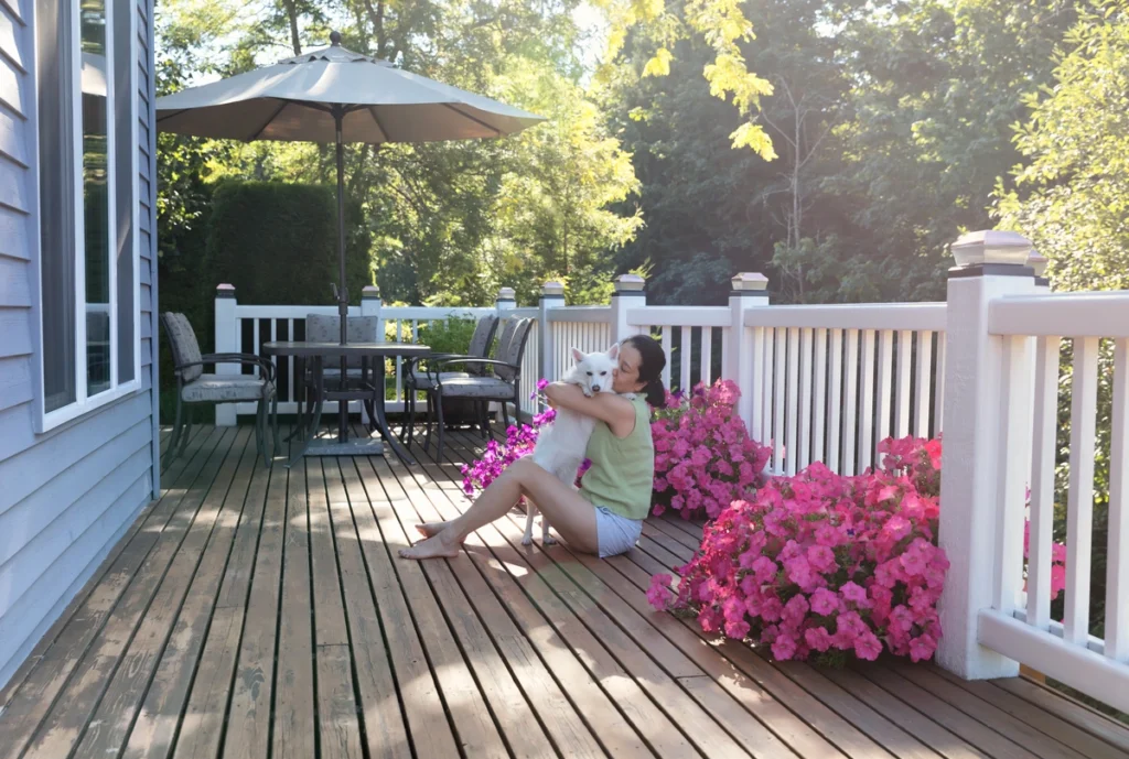 A woman sits on a wooden deck made of the best decking material, hugging a small white dog. Bright pink flowers in planters line the railing, and a patio table with an umbrella is in the background. Lush green trees surround the sunlit area.