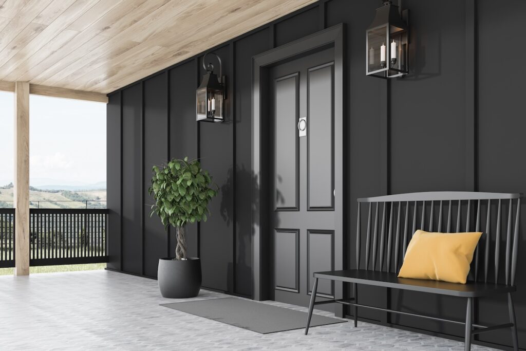 A modern porch with a black door and lanterns on both sides. A potted plant sits near the door. A black bench with a yellow cushion is on the right. The space features wood and metal accents, overlooking a distant landscape.