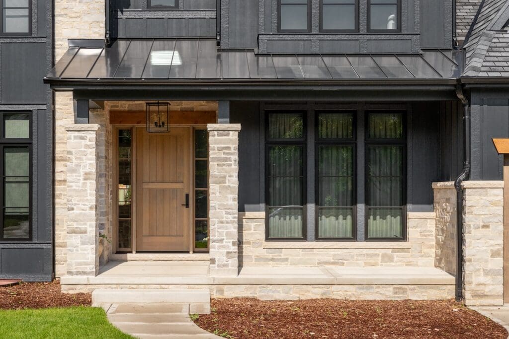 Front entrance of a modern house featuring a wooden door with a black handle, light stone walls, and black-framed windows. The porch is supported by stone columns and a path leads to the door through a landscaped area.