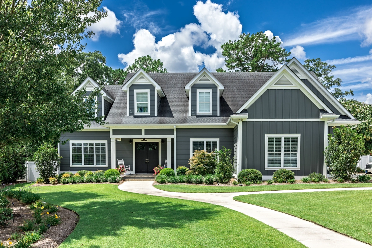 A charming gray house with siding, featuring a gabled roof and dormer windows, sits behind a manicured lawn in Mahtomedi. A winding concrete path leads to the entrance, flanked by lush greenery and trees. A clear blue sky with fluffy clouds frames the scene.