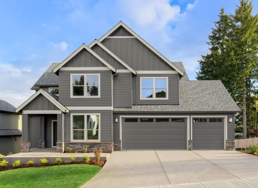 A modern, two-story gray house with diverse siding options stands gracefully amid a neatly landscaped front yard. It features a three-car garage, large windows, and a gabled roof. Tall trees are visible in the background under a partly cloudy sky.