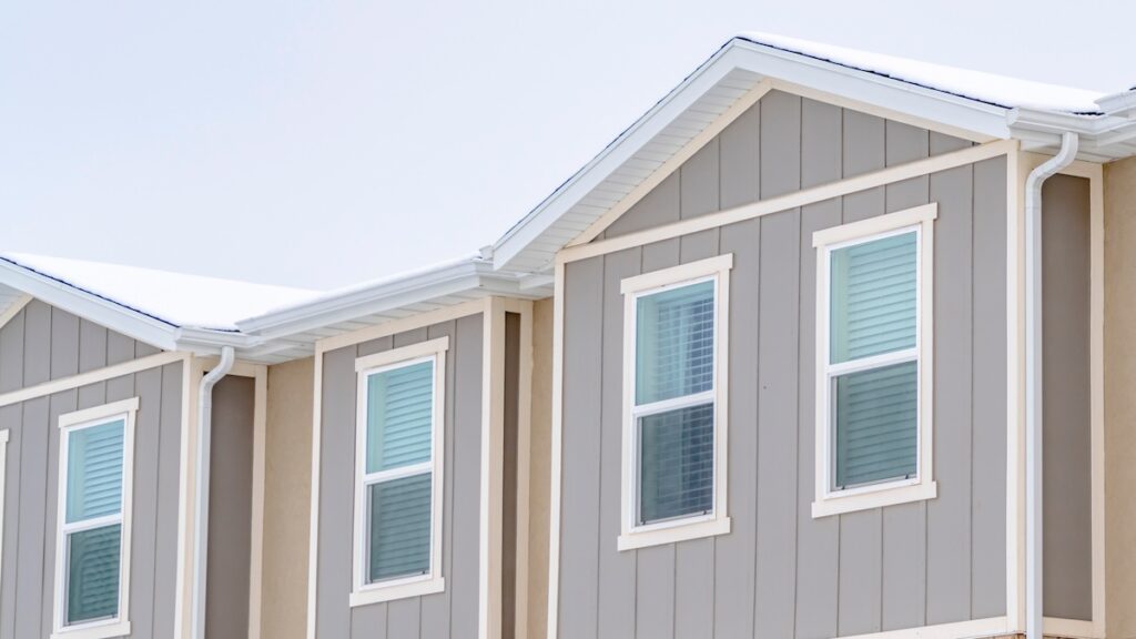 The image shows the upper section of a modern residential building with light gray siding and white trim. There are multiple rectangular windows with blue tint. The roof has a gentle slope and is covered with a thin layer of snow.