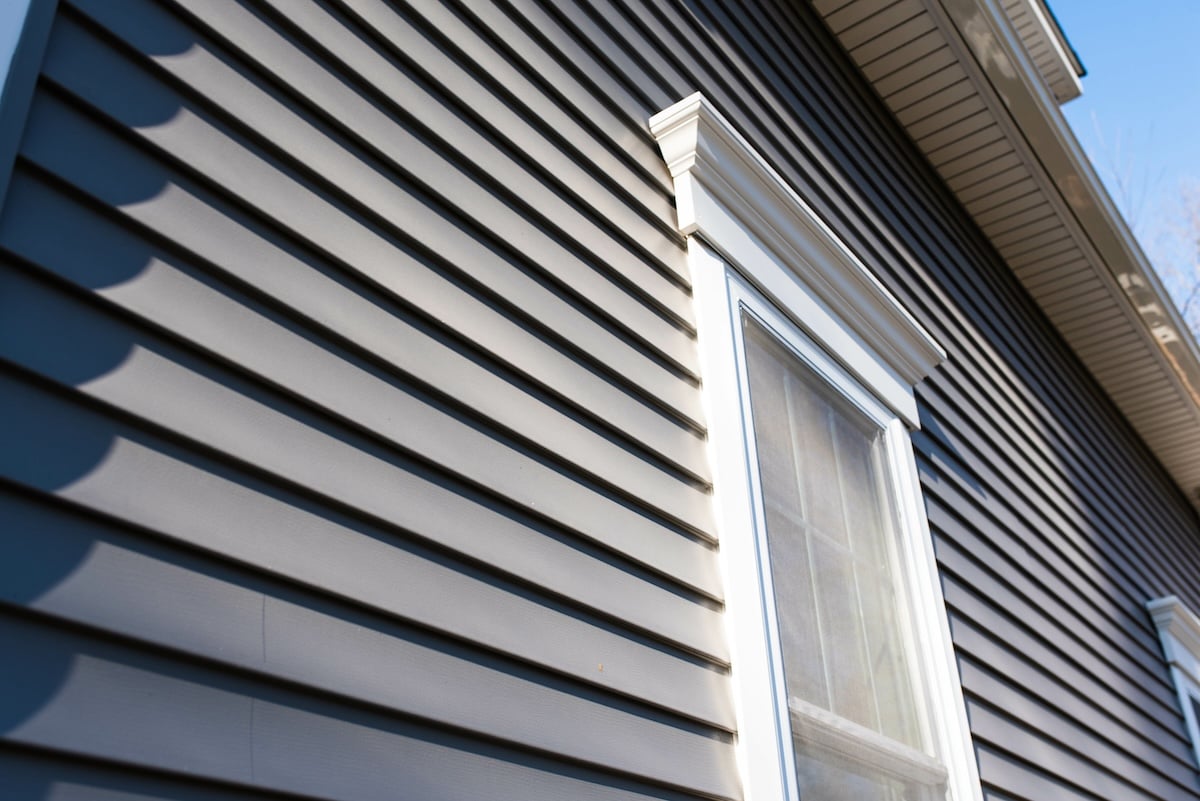 A close-up angle of a house with gray vinyl siding and a white-trimmed window. The siding features horizontal panels, and sunlight casts shadows across the surface. The roof's eaves are visible at the top edge of the image.