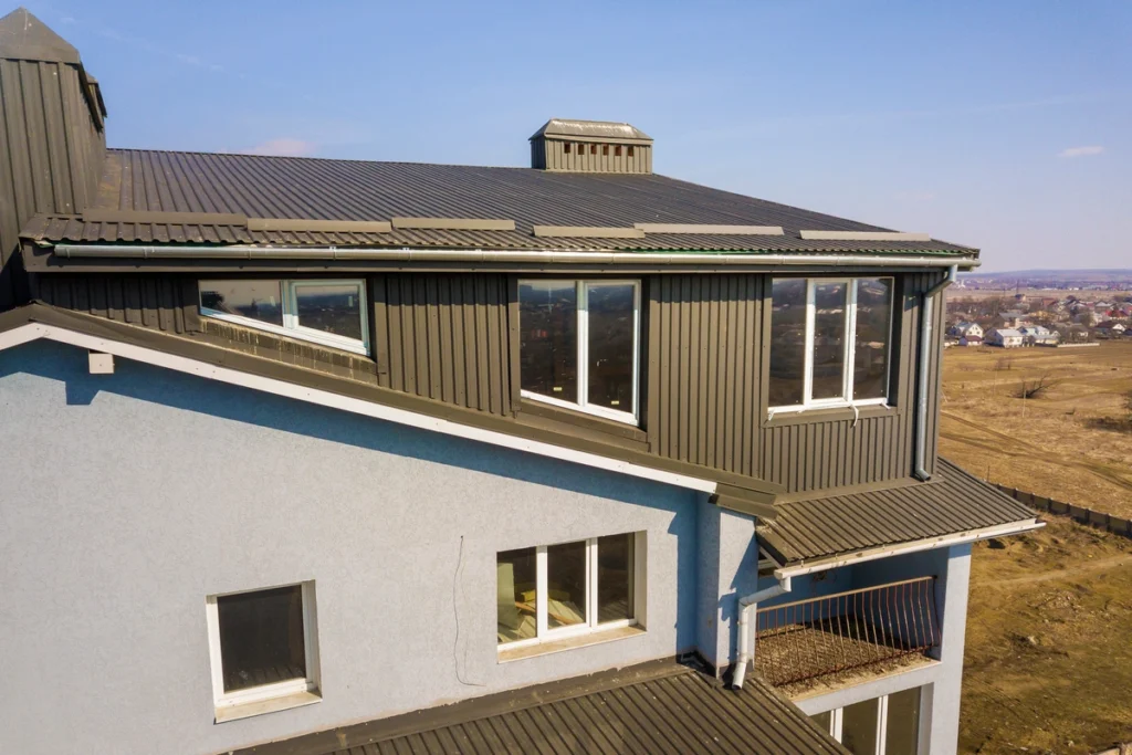 A two-story building with a light blue facade and sleek house siding options features dark metal roof panels. The upper level boasts several large windows, offering views of a rural landscape with fields and distant houses under a clear blue sky.