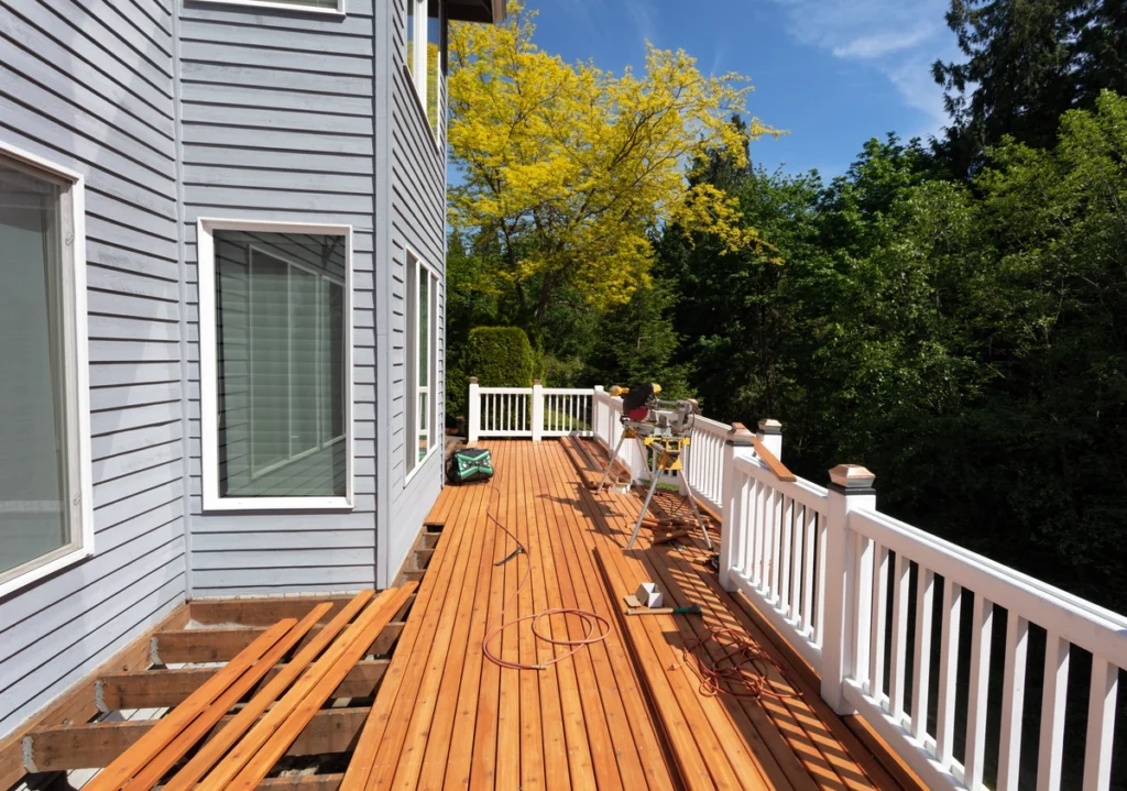 A newly constructed wooden deck, made with the best decking material, is attached to a light blue house with white railings. Construction tools and materials are scattered on the deck. Lush green trees and a bright yellow tree stand in the background under a clear blue sky.