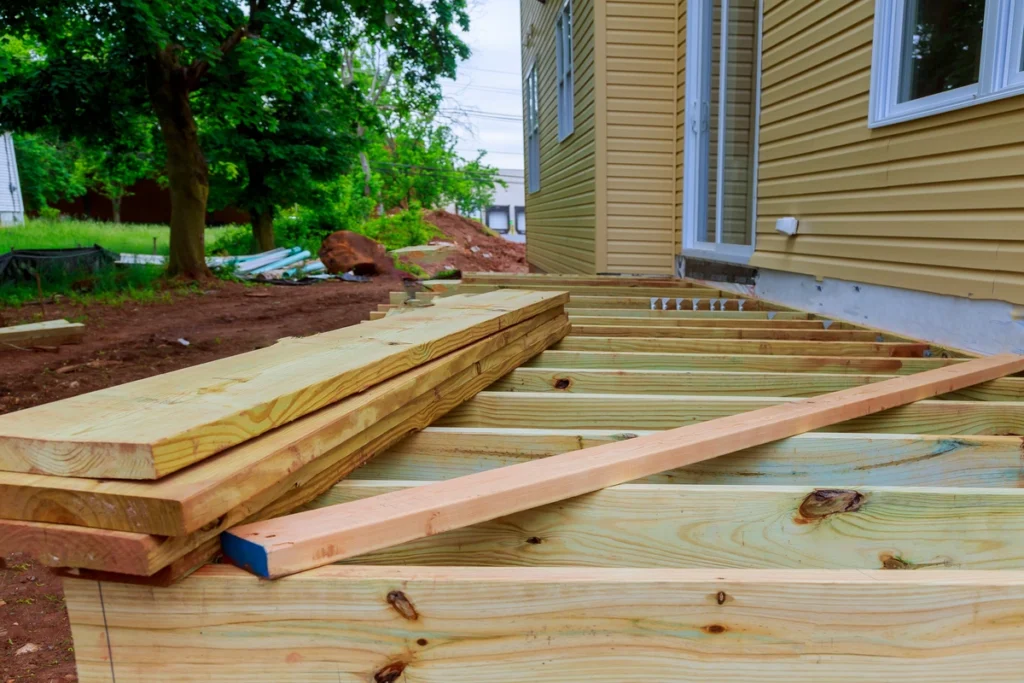 Lumber is strategically placed on a deck under construction, attached to a beige house. Piles of wooden planks await their turn on the unfinished deck as tools and materials for learning how to replace deck boards lie scattered around. A green tree stands serenely in the background alongside earthy soil.