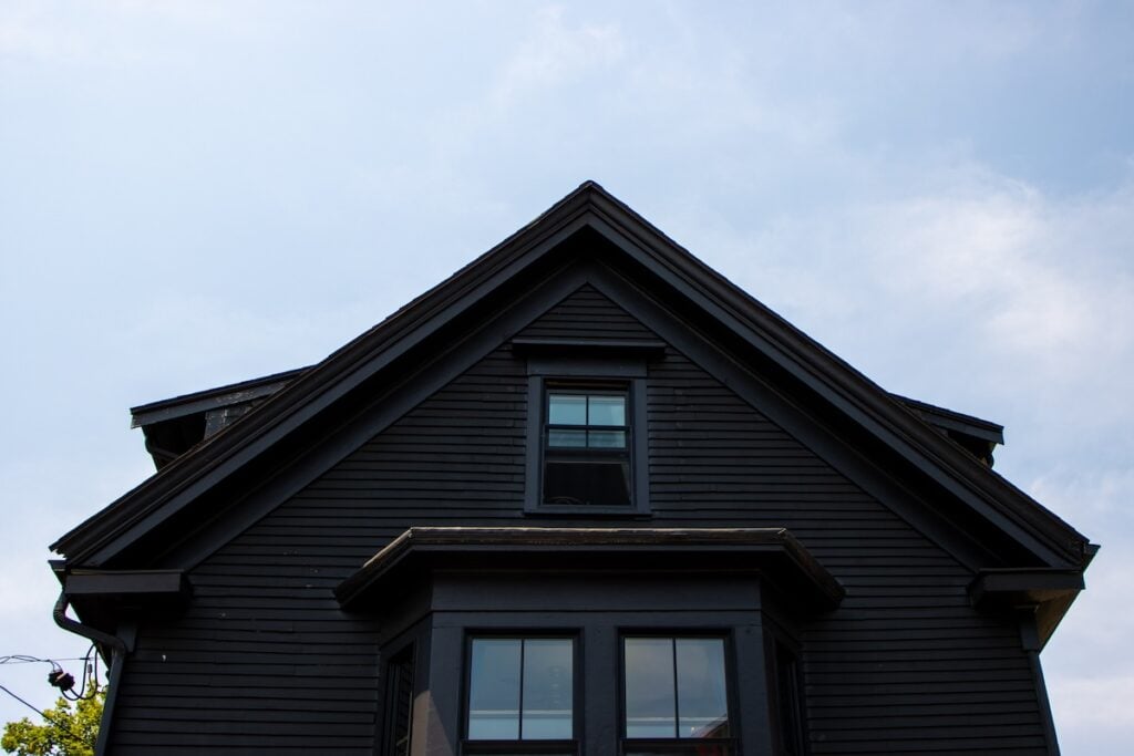 A dark grey house with a triangular roof and two windows is pictured against a clear blue sky. The front facade has wood paneling and a small overhang above the ground-level window. Some green foliage is partially visible on the left side.