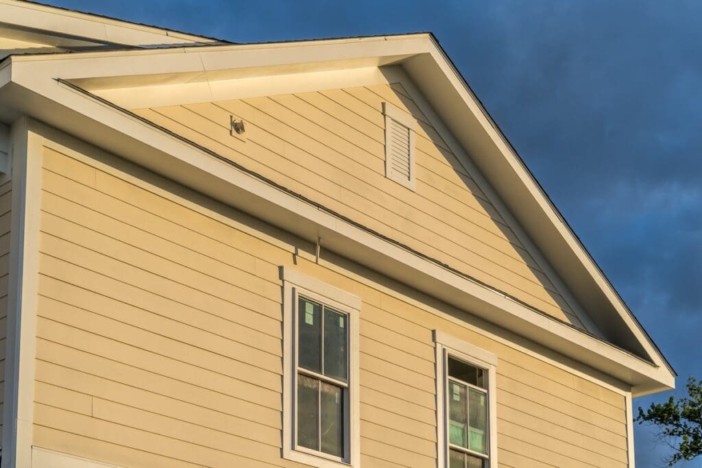 A beige house with horizontal siding is shown under a blue sky. It has two windows with white frames on the upper level and a small vent near the roof peak. The lighting suggests it's either early morning or late afternoon.