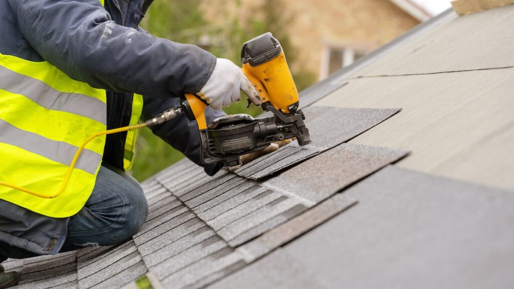 Person wearing a high-visibility jacket and gloves, kneeling, using a nail gun to install asphalt shingles on a sloped roof.