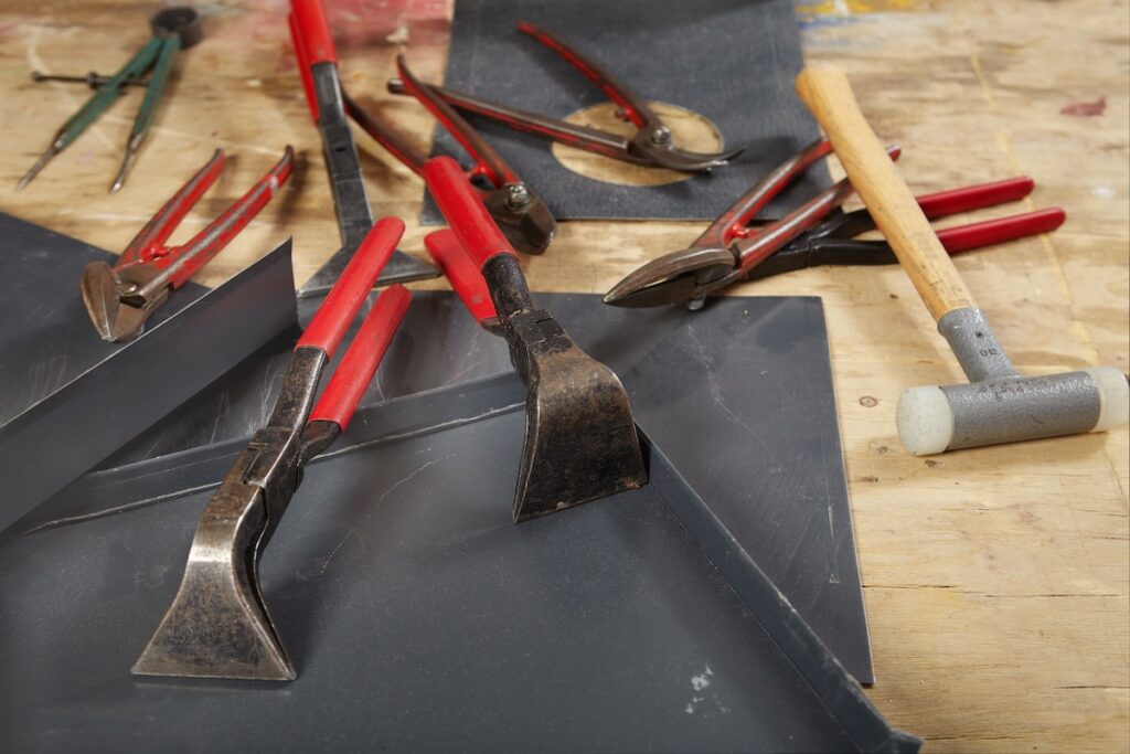 A workbench with various tools, including red-handled pliers, a compass, a wrench, and a hammer with a light-colored head. Gray metal sheets are also present, suggesting a metalworking project in progress.