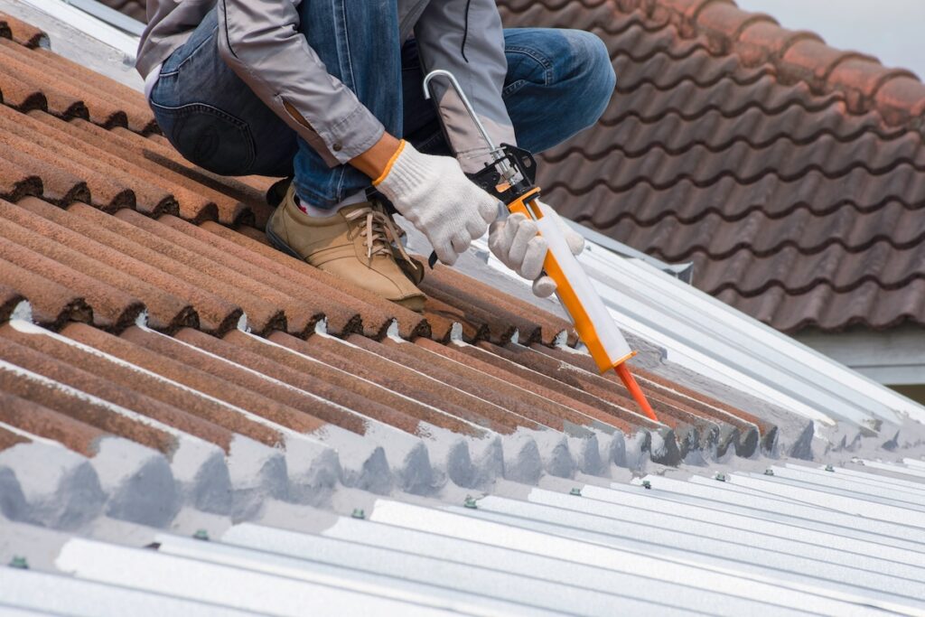 A person wearing gloves and casual shoes is kneeling on a roof, using a caulking gun to seal gaps between corrugated metal sheets. The roof features both red tiles and silver metal panels.