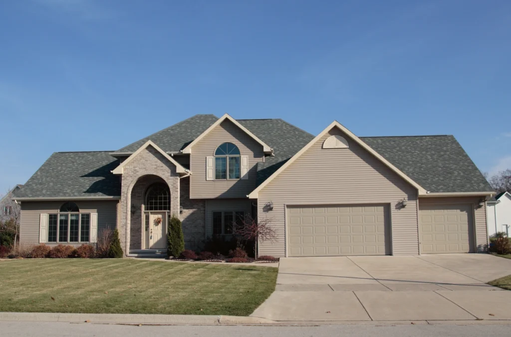 A two-story suburban house with a large driveway and attached three-car garage boasts diverse house siding options. The home features a brick archway entrance, several windows, and a green lawn under the clear blue sky.