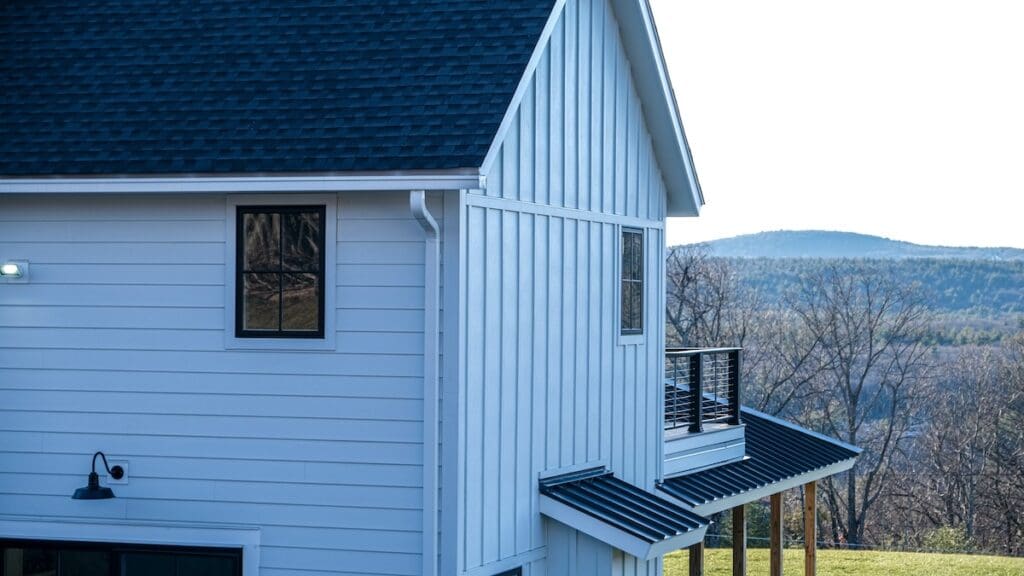 A modern white house with blue trim and a balcony, set against a backdrop of rolling hills and leafless trees. The sky is clear, adding to the serene rural landscape.