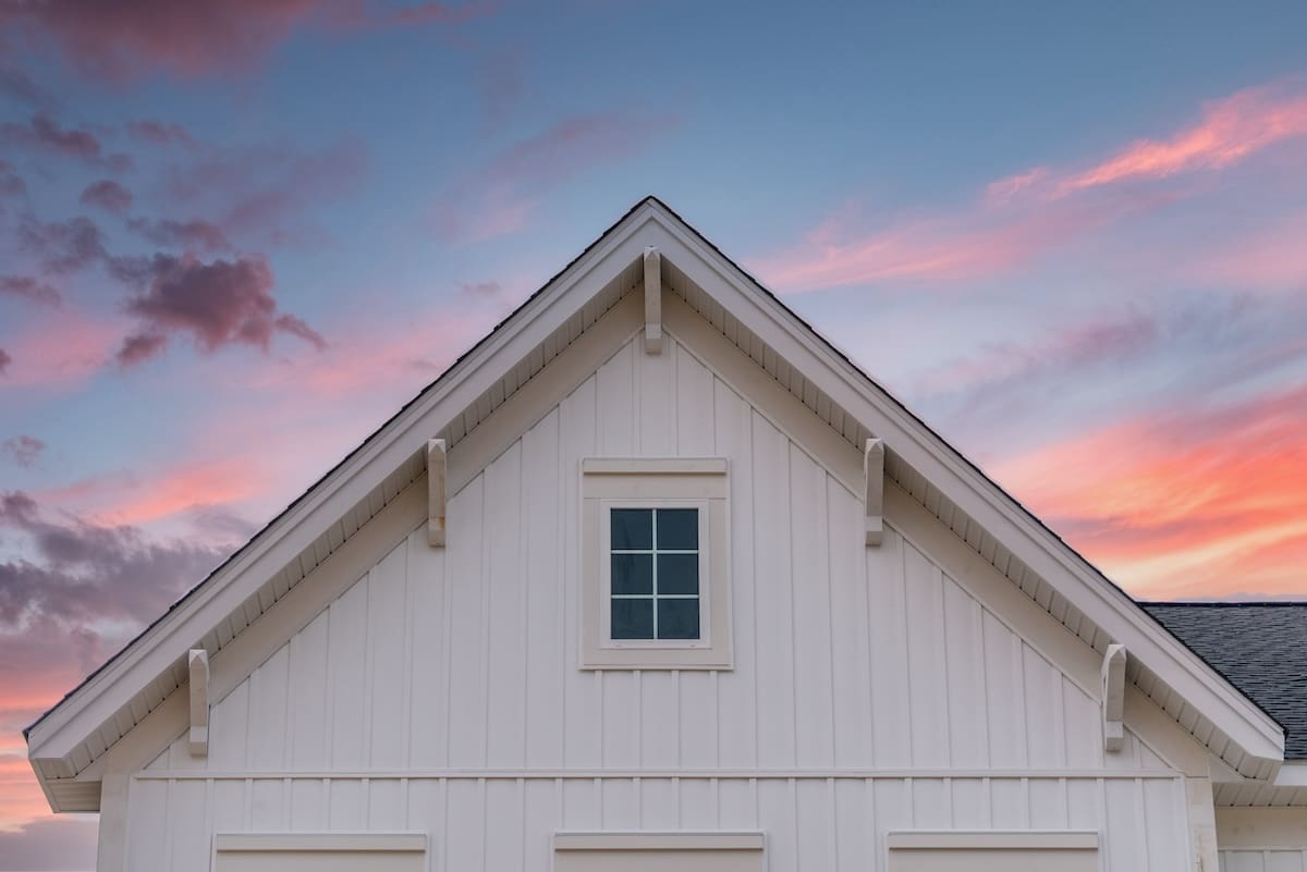 White vertical vinyl siding gable with decorative brackets and attic window against sunset sky