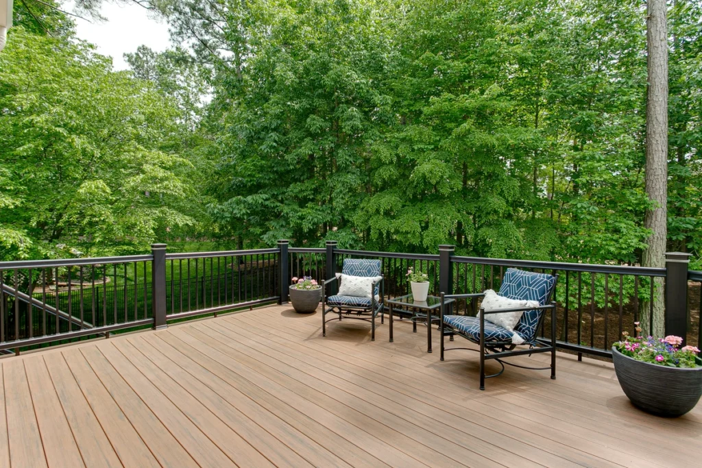 A wooden deck, made from the best decking material, features two cushioned chairs and a small glass table set amidst lush green trees. Three potted plants with flowers line the railing, enhancing the natural backdrop of dense foliage.