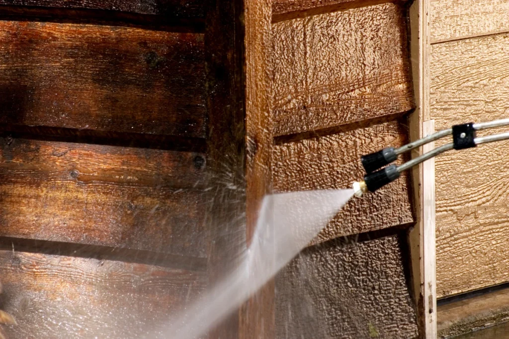 A close-up of a pressure washer cleaning a wooden surface demonstrates how to clean wood siding effectively. The water spray visibly removes dirt, revealing a lighter shade. Two nozzles are seen at the end of the pressure washer, ensuring precise and thorough cleaning.