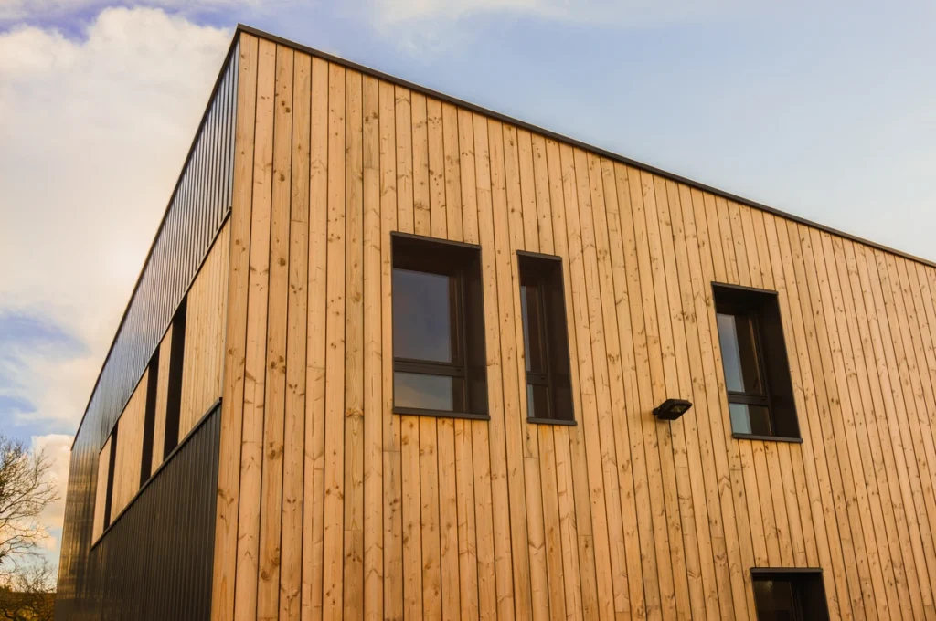 Modern building with a facade of vertical wooden planks and contrasting dark panels. The clean wood siding complements the large windows, all under a clear blue sky with soft clouds.