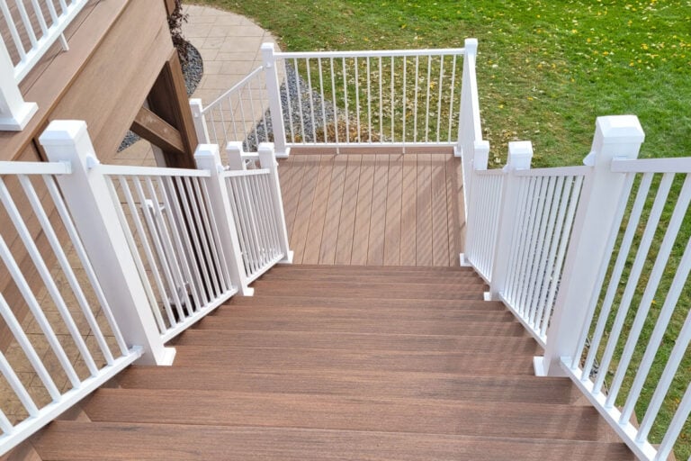 A downward view of brown wooden deck stairs with white railings, leading to a small landing and then to a grassy backyard area.