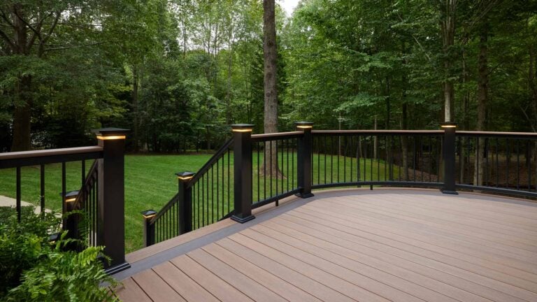 A spacious wooden deck with black railings and post lights overlooks a lush, green backyard surrounded by tall trees. Steps lead from the deck down to the grassy lawn.