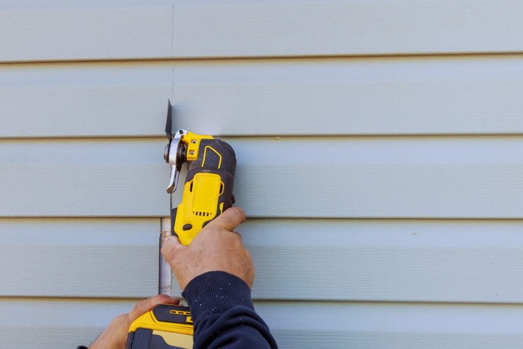 Person using a yellow and black oscillating multi-tool to cut into light blue vinyl siding on a house.