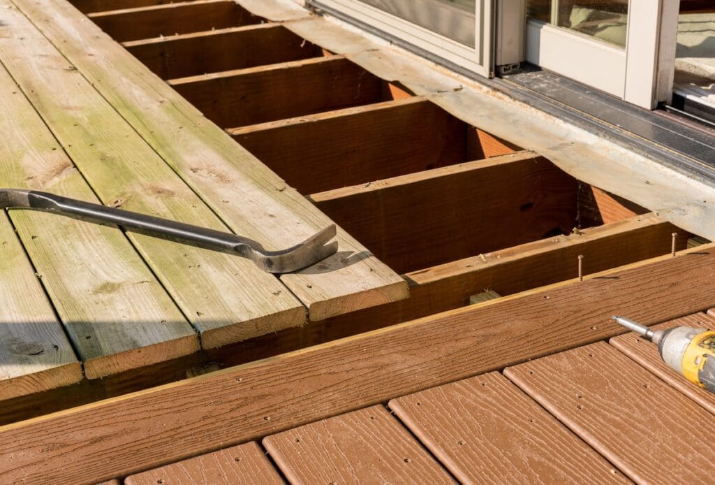 A deck under renovation shows old, weathered wooden planks being replaced with new brown composite boards