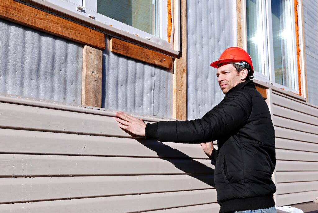A man wearing a red hard hat and black jacket inspects beige siding on a building.
