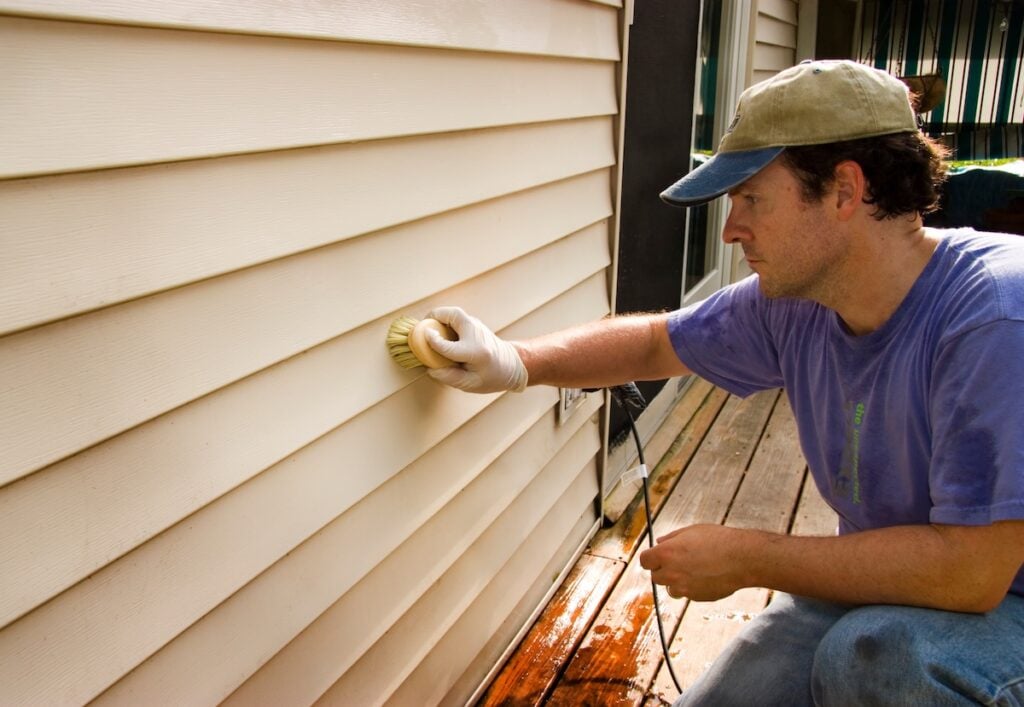 Person wearing a cap and gloves scrubbing the siding of a house with a brush.
