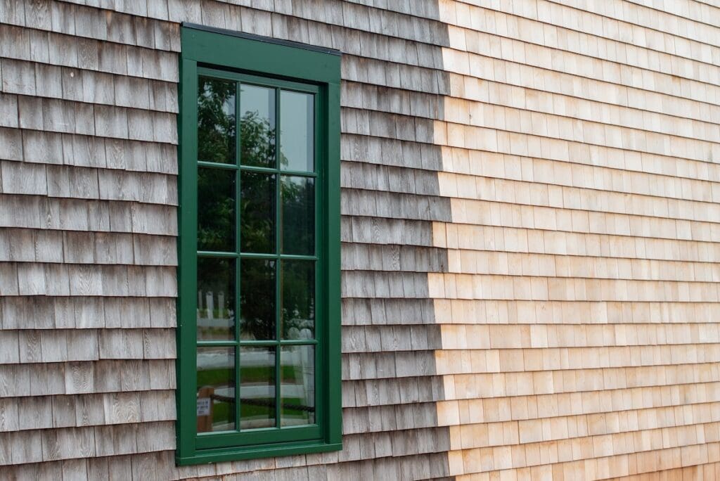 A green-framed window is set in a wooden shingle wall of cedar siding, with the left side weathered gray and the right side fresh and light-colored.