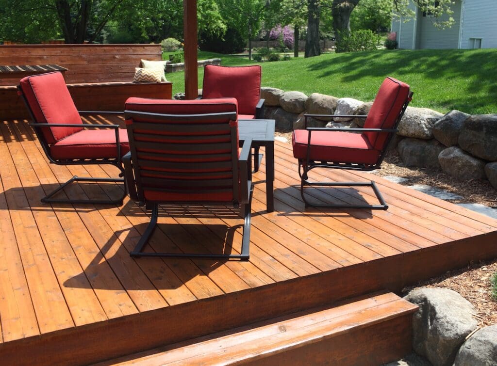 Four red cushioned patio chairs surround a small square table on cedar decking boards, framed by a stone border and lush green lawn in the background.