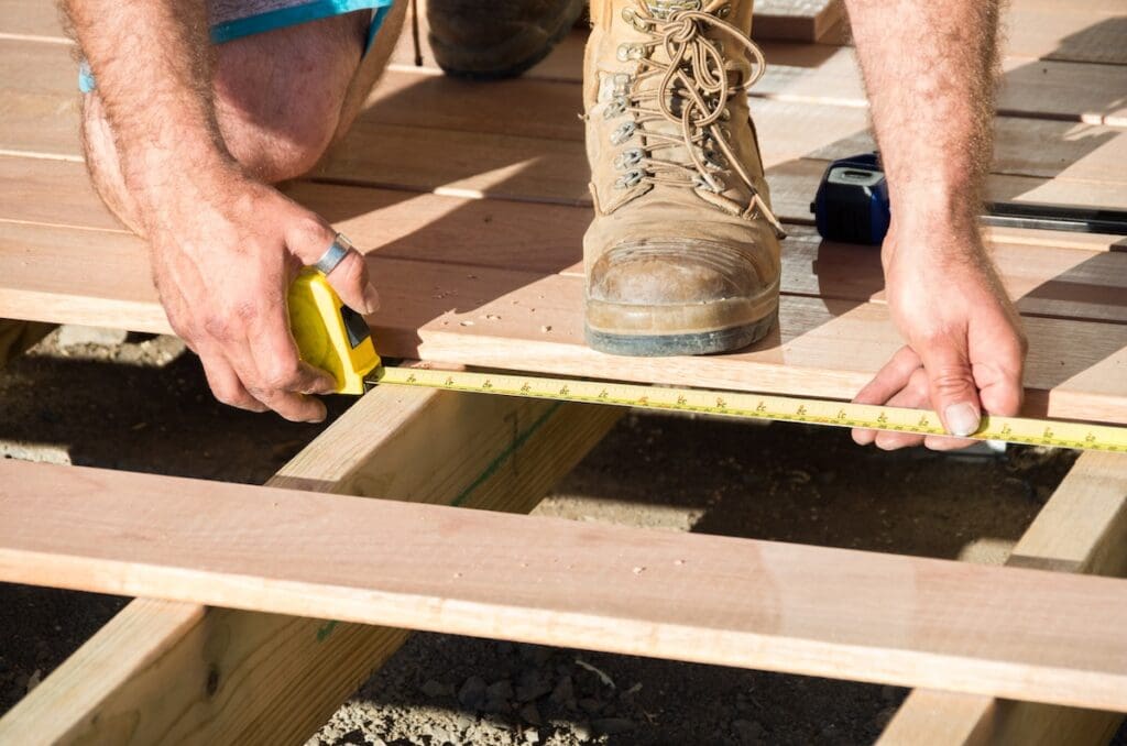 A person wearing work boots measures a wooden plank with a yellow tape measure on an outdoor deck under construction