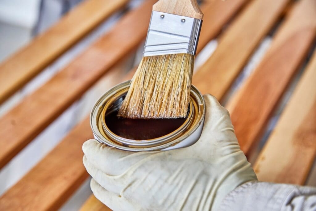 A gloved hand holds a can of brown wood stain while a paintbrush is dipped into it