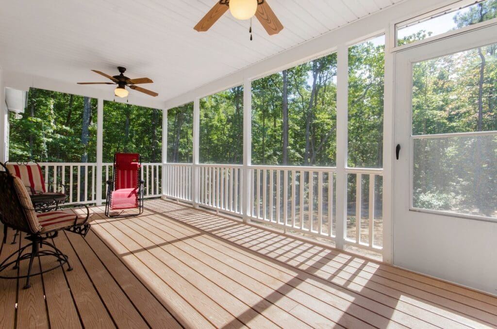 A screened-in deck with wooden flooring, white railings, two ceiling fans, a red lounge chair
