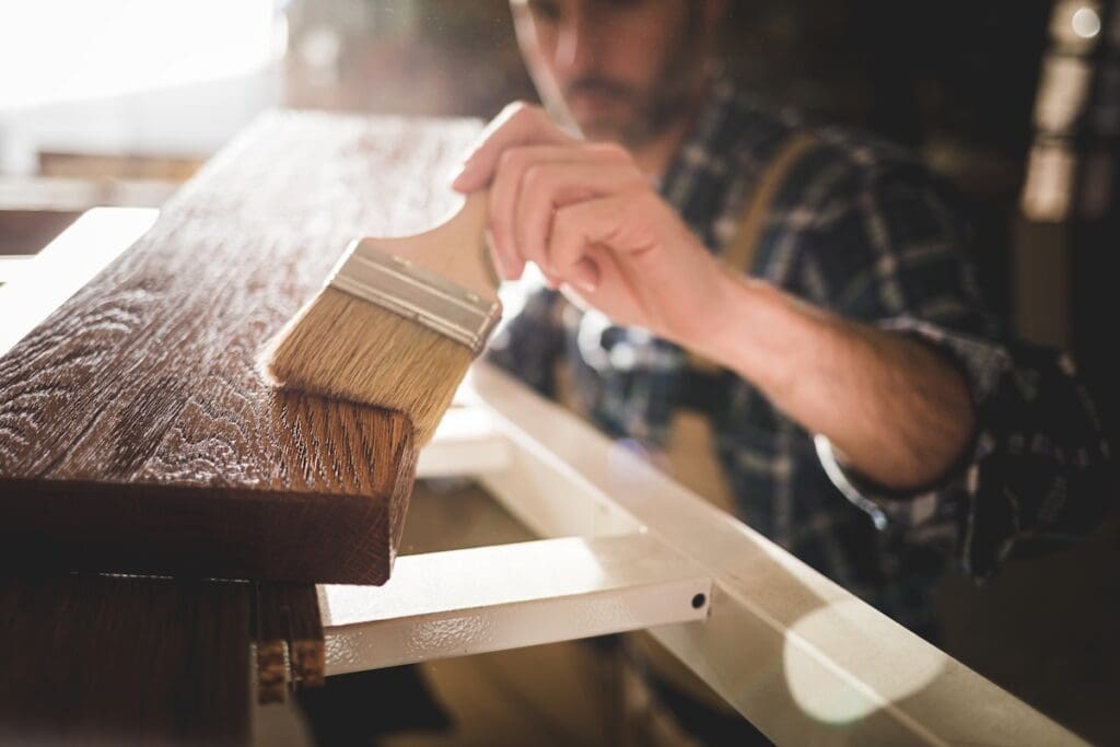 A person in a plaid shirt and apron applies varnish to a wooden plank with a wide paintbrush