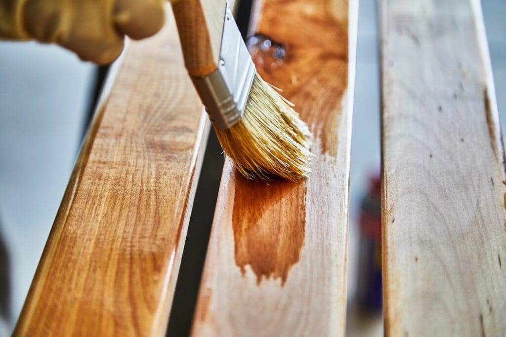 A close-up of a hand holding a paintbrush applying varnish to a wooden surface