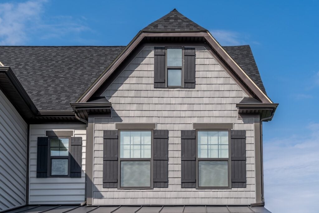 The image shows the upper part of a house exterior with gray shaker siding, black roof shingles, and four windows featuring black shutters against a blue sky.