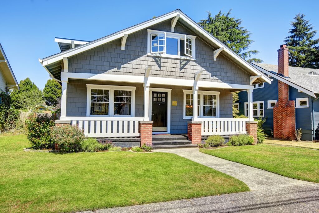 A charming grey Craftsman-style house with a covered front porch, white trim, and brick pillars sits on a well-manicured lawn