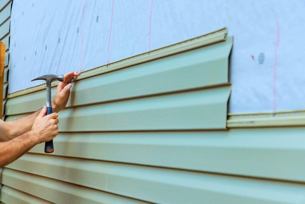 A person uses a hammer to install light green vinyl siding panels on the exterior of a house, over a white house wrap.