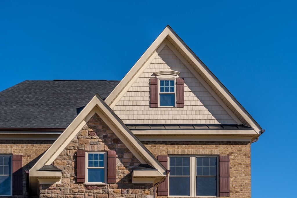 A close-up of a house featuring stone and brick walls, shaker siding, multiple gables, and several windows with brown shutters