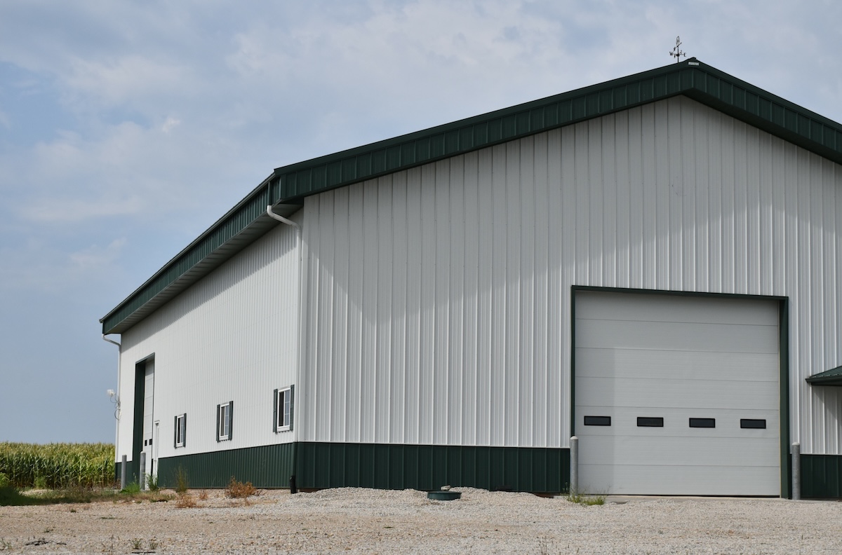 A large white metal building with low maintenance siding and a dark green roof and trim, featuring a large garage door and several small windows, sits on a gravel lot with a cornfield in the background under a partly cloudy sky.