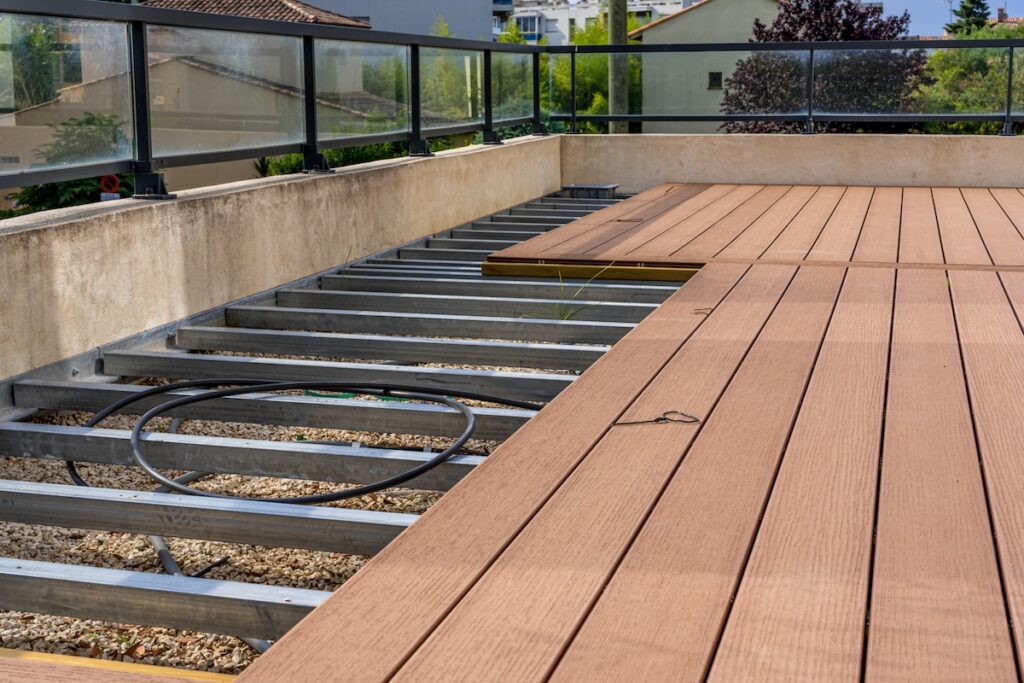 A partially constructed rooftop deck, combining composite deck installation with exposed metal framing and gravel underneath, features some boards already in place and city buildings in the background.