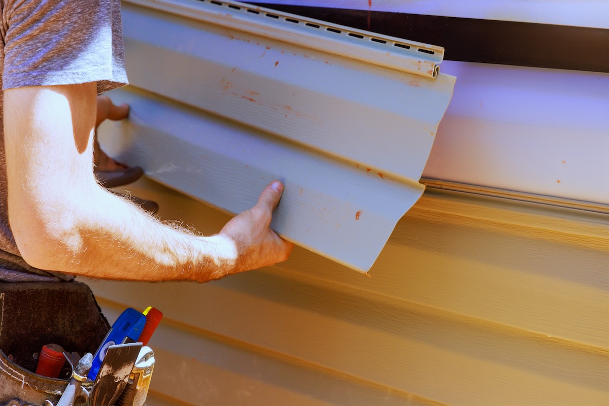 A person installs beige vinyl siding on a house, carefully holding a panel in place to avoid warped siding, while wearing a tool belt with various tools visible.