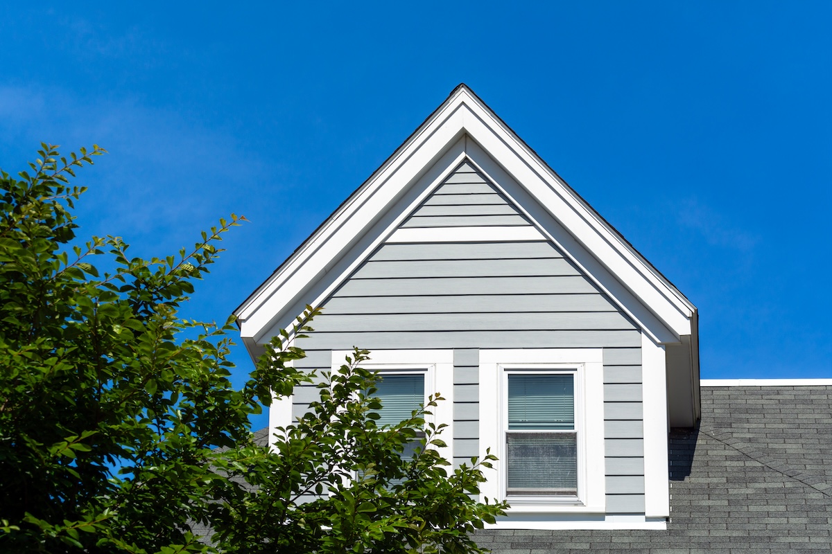 A gray house with white trim siding features two windows beneath a pointed roof, set against a clear blue sky with green tree branches in the foreground.